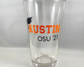 May include: A clear glass pint glass with an orange and black design. The design says "Austin OSU '21".