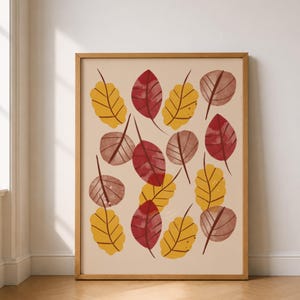 Könnte beinhalten: Ein rechteckiger Wandkunstdruck in einem hellen Holzrahmen mit einem Muster aus verstreuten Herbstblättern in Rot-, Gelb- und gedämpften Brauntönen auf beigem Hintergrund. Die Blätter weisen deutlich sichtbare dunkelbraune Blattadern auf.