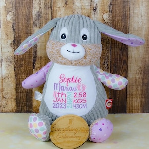May include: Grey plush bunny with pink and purple fabric accents. The bunny has a white embroidered patch with the text "Sophie Marie" and "11th JAN 2023 2:18PM 2.58 KGS 43CM". A wooden tag with the text "Everything Nice Embroidery" is in front of the bunny.