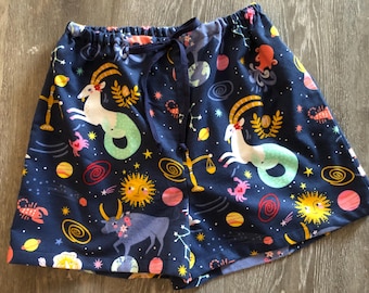 Könnte beinhalten: Dunkelblaue Shorts mit einem Allover-Print von Tierkreiszeichen, Planeten und Sternen. Die Shorts haben einen Kordelzug in der Taille und eine Vielzahl von farbenfrohen astrologischen Symbolen, darunter eine Sonne, ein Stier und ein Ziegen-Meerjungfrau-Hybrid.
