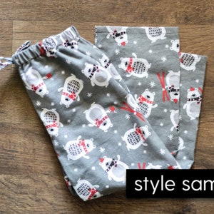 May include: Gray pajama pants with a repeating pattern of white polar bears wearing red scarves and hats. The bears are interspersed with red skis and white snowflakes. The pants have a drawstring waist and the words "style sample" in a black rectangle.