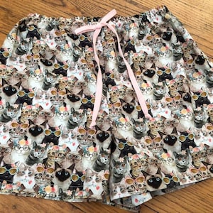 May include: A pair of patterned shorts featuring a repeating design of cats wearing sunglasses and glasses. The shorts have a pink drawstring and are made of a lightweight fabric. The cats are in various colors and poses.