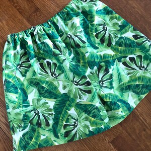 May include: A skirt with an elasticated waistband, displaying a vivid tropical leaf pattern in green and white. The fabric is adorned with large banana leaves and other foliage, creating a summery, botanical design.