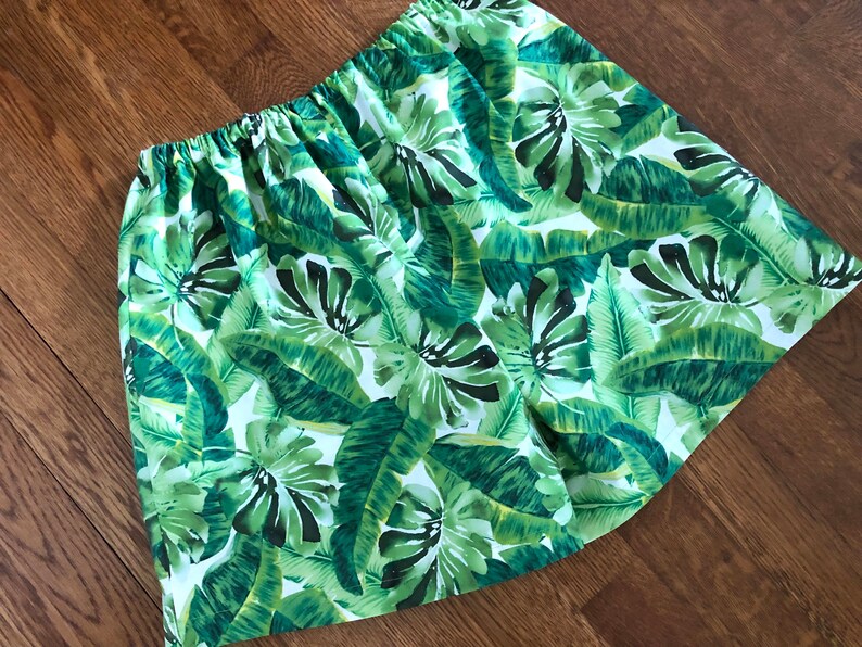 May include: A skirt with an elasticated waistband, displaying a vivid tropical leaf pattern in green and white. The fabric is adorned with large banana leaves and other foliage, creating a summery, botanical design.