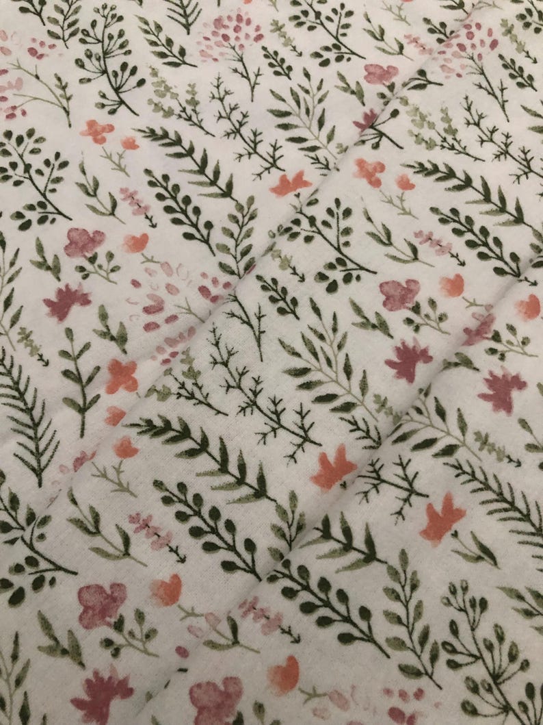 Puede incluir: Tela blanca con un estampado floral con hojas verdes y flores rosas y melocot&oacute;n. El dise&ntilde;o es un patr&oacute;n repetitivo de elementos bot&aacute;nicos, creando una est&eacute;tica suave y natural. La tela tiene una textura acolchada.