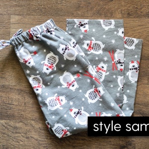 Puede incluir: Pantalones de pijama grises con un estampado repetido de osos polares blancos con bufandas y gorros rojos, con copos de nieve y gr&aacute;ficos de esqu&iacute; rojos. Los pantalones tienen cintura con cord&oacute;n y el texto "style sample".