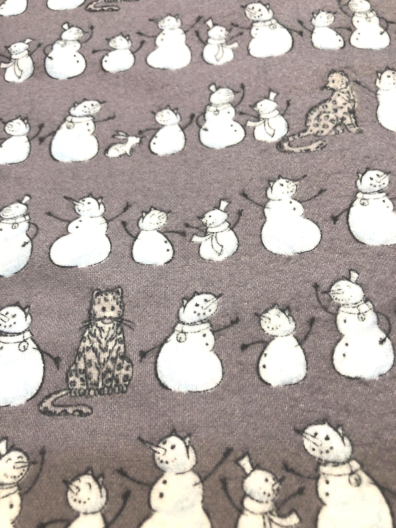 K&ouml;nnte beinhalten: Ein grauer Stoff mit einem sich wiederholenden Muster aus wei&szlig;en Schneem&auml;nnern mit schwarzen Stockarmen und Karottennasen. Dazwischen befinden sich Illustrationen von Katzen mit leopardenartigen Flecken. Die Schneem&auml;nner haben verschiedene Accessoires.