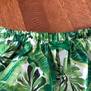 May include: A close-up of a skirt with a tropical leaf pattern in shades of green and white. The waistband is elasticated and has a small green bow. The fabric is lightweight.