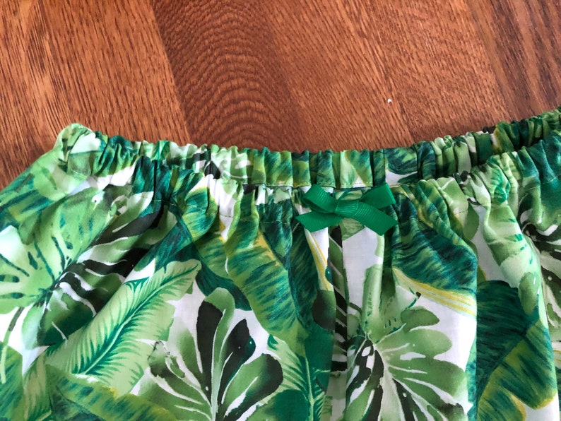 May include: A close-up of a skirt with a tropical leaf pattern in shades of green and white. The waistband is elasticated and has a small green bow. The fabric is lightweight.