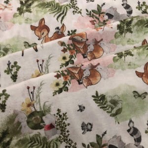 May include: White fabric with a repeating pattern of woodland creatures and floral elements. The design features deer, foxes, raccoons, and rabbits amidst flowers and greenery in soft pastel colors. The overall theme is a whimsical forest scene.