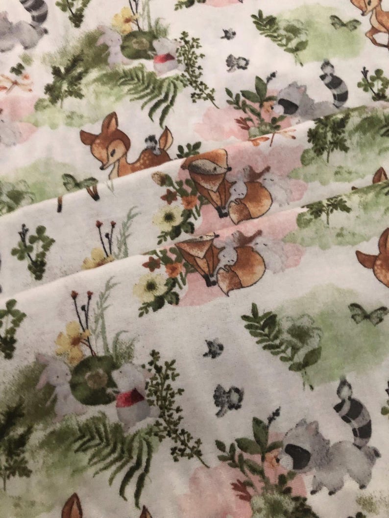 May include: White fabric with a repeating pattern of woodland creatures and floral elements. The design features deer, foxes, raccoons, and rabbits amidst flowers and greenery in soft pastel colors. The overall theme is a whimsical forest scene.