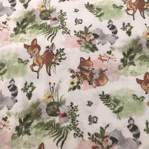 May include: White fabric with a repeating pattern of woodland animals and floral elements. The design features deer, foxes, raccoons, and rabbits, along with flowers, butterflies, and greenery in soft colors.