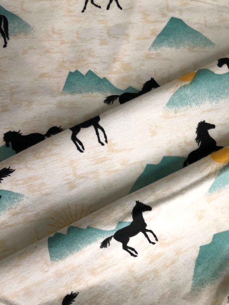 May include: Cream-colored fabric with a repeating pattern of black horses, teal mountains, and yellow suns. The horses are in various poses, some galloping, others standing. The design has a rustic, outdoorsy feel.