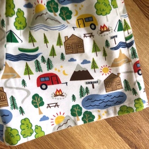 May include: White fabric with a repeating pattern of camping scenes. The pattern includes cabins, trees, mountains, lakes, campfires, and stars. The fabric has a blue, green, red, yellow, and brown colour scheme.