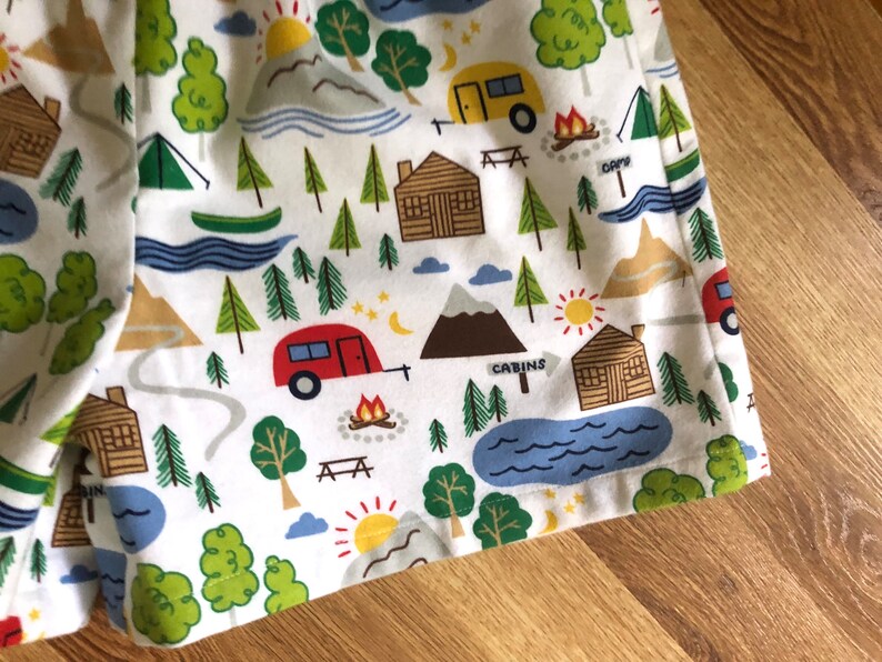 May include: White fabric with a repeating pattern of camping scenes. The pattern includes cabins, trees, mountains, lakes, campfires, and stars. The fabric has a blue, green, red, yellow, and brown colour scheme.