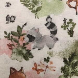 May include: White fabric with a repeating pattern of woodland creatures and floral elements. The design includes raccoons, foxes, deer, butterflies, and green foliage, set against soft pink and green watercolor-style backgrounds.