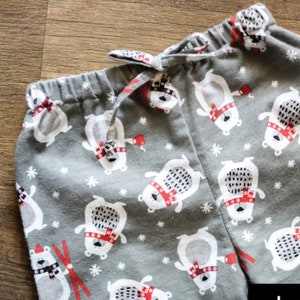 May include: Gray pajama pants with a repeating pattern of white polar bears wearing red scarves and hats, and red mittens. The pants have a drawstring waist and white snowflakes scattered throughout the design.