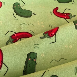 May include: Green and red fabric with a repeating pattern of smiling green pickles and red chilli peppers. The pickles and peppers are cartoon-like and have arms and legs. The fabric has a light green background.