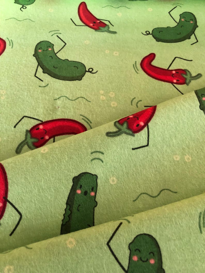 May include: Green and red fabric with a repeating pattern of smiling green pickles and red chilli peppers. The pickles and peppers are cartoon-like and have arms and legs. The fabric has a light green background.