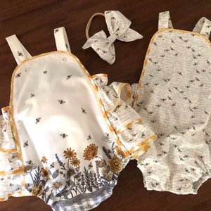 May include: Two baby onesies with a bee theme. One onesie is white with a bee print and a floral border. The other onesie is grey with a bee print. Both onesies have yellow trim.