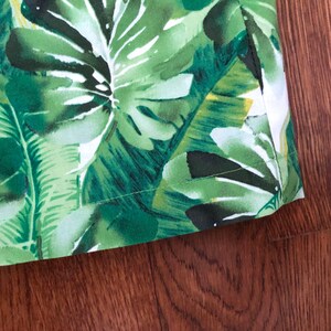 May include: Close-up of a fabric with a vibrant tropical leaf pattern in shades of green and white. The fabric features large, detailed leaves, creating a lush, botanical design. The edge of the fabric is visible, showing a neat seam.