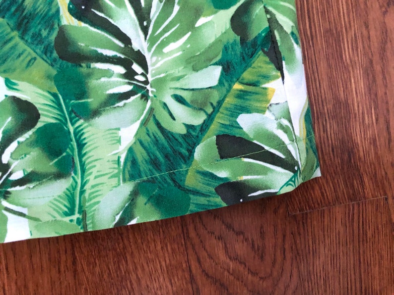 May include: Close-up of a fabric with a vibrant tropical leaf pattern in shades of green and white. The fabric features large, detailed leaves, creating a lush, botanical design. The edge of the fabric is visible, showing a neat seam.