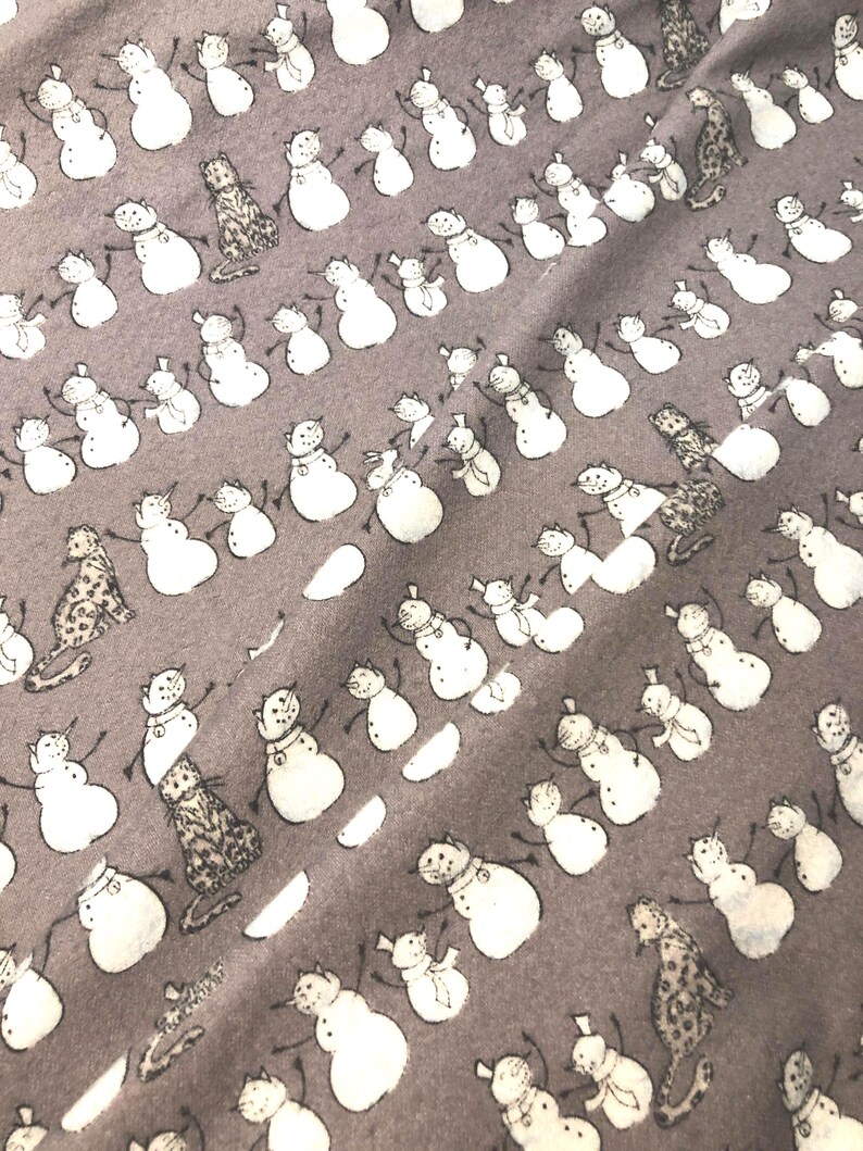 K&ouml;nnte beinhalten: Ein grauer Stoff mit einem sich wiederholenden Muster aus wei&szlig;en Schneem&auml;nnern und Leopardenfiguren. Die Schneem&auml;nner haben schwarze Arme und Karottennasen. Der Stoff hat ein weiches, leicht strukturiertes Aussehen.