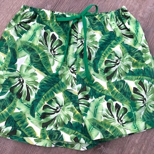 May include: Shorts with a tropical leaf print in green and white, featuring a green drawstring. The elasticated waistband and lightweight fabric suggest a comfortable fit. The shorts are patterned with large leaves.