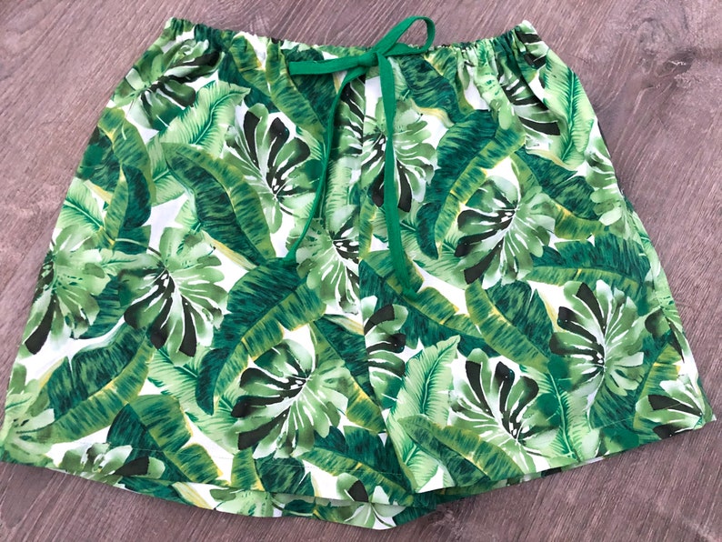 May include: Shorts with a tropical leaf print in green and white, featuring a green drawstring. The elasticated waistband and lightweight fabric suggest a comfortable fit. The shorts are patterned with large leaves.