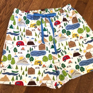 May include: White cotton shorts with a blue drawstring and a repeating pattern of camping scenes, including cabins, mountains, trees, and campers.
