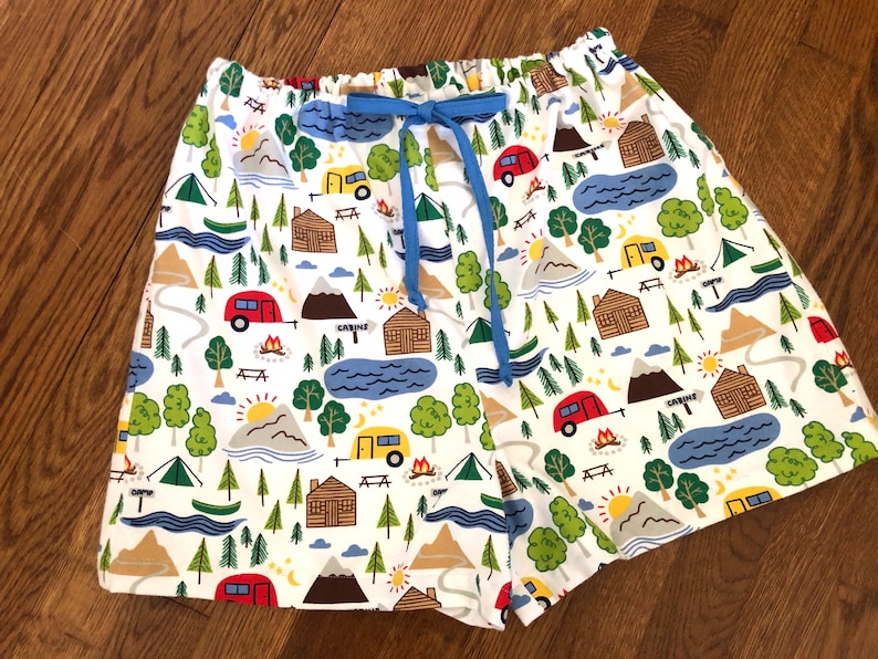 May include: White cotton shorts with a blue drawstring and a repeating pattern of camping scenes, including cabins, mountains, trees, and campers.