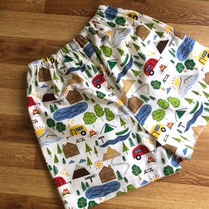 May include: White cotton fabric with a repeating pattern of camping-themed illustrations. The pattern includes red, yellow, blue, and green camper vans, cabins, mountains, trees, and campfires.