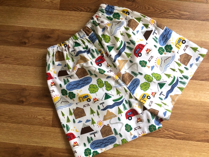 May include: White cotton fabric with a repeating pattern of camping-themed illustrations. The pattern includes red, yellow, blue, and green camper vans, cabins, mountains, trees, and campfires.