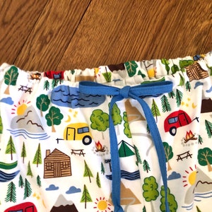 May include: White fabric with a camping themed print featuring cabins, trees, mountains, lakes, and campers. The fabric has a blue drawstring.