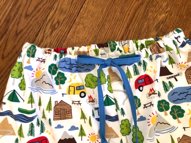 May include: White fabric with a camping themed print featuring cabins, trees, mountains, lakes, and campers. The fabric has a blue drawstring.