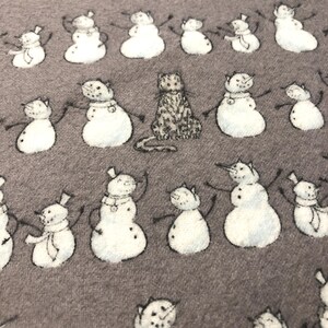 K&ouml;nnte beinhalten: Ein grauer Stoff mit einem sich wiederholenden Muster aus Schneem&auml;nnern mit Katzenk&ouml;pfen und einer einzelnen Katze. Die Schneem&auml;nner haben schwarze St&auml;bchenarme und tragen H&uuml;te und Schals. Die Katze sitzt aufrecht.