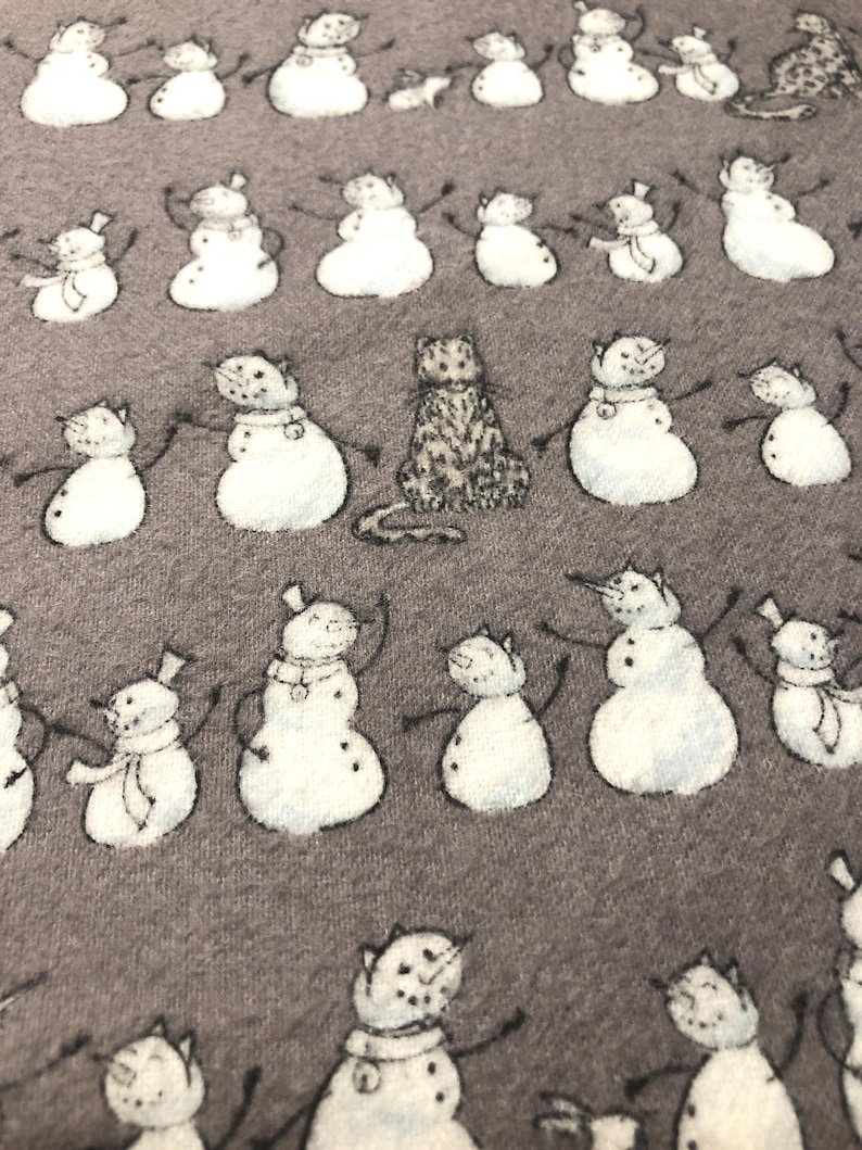 K&ouml;nnte beinhalten: Ein grauer Stoff mit einem sich wiederholenden Muster aus Schneem&auml;nnern mit Katzenk&ouml;pfen und einer einzelnen Katze. Die Schneem&auml;nner haben schwarze St&auml;bchenarme und tragen H&uuml;te und Schals. Die Katze sitzt aufrecht.