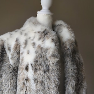 May include: A long-sleeved coat made of faux fur featuring a white base color with mottled grey and brown spots throughout the fabric. The coat has a wide collar and is displayed on a white mannequin torso against a plain, neutral-colored background.