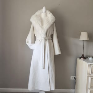 Könnte beinhalten: Ein langer, cremeweißer Mantel mit einem breiten, flauschigen Kunstfellkragen, der sich über die Vorderseite erstreckt. Der Mantel verfügt über lange, weite Ärmel und einen passenden Bindegürtel aus Stoff in der Taille für eine strukturierte Form.