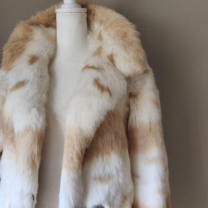 May include: A long-sleeved coat featuring a thick, plush texture with a variegated pattern of white and light tan fur. The garment has a wide, prominent collar and a double-breasted front closure with large, circular, metallic-toned buttons.
