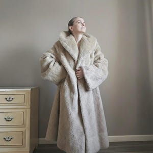 Op de afbeelding: Een lange, beige imitatiebontjas met een brede kraag en grote mouwen. De jas is vloerlang en ziet er zacht en pluche uit. De persoon die de jas draagt, kijkt omhoog. Een beige dressoir is zichtbaar op de achtergrond.