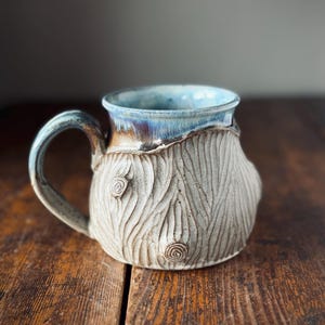 Peut inclure: Une tasse en céramique avec un extérieur texturé blanc cassé présentant un motif de grain de bois sculpté et deux nœuds visibles. Le bord supérieur et l'anse sont recouverts d'un vernis brillant bleu et brun irisé qui coule sur le côté.