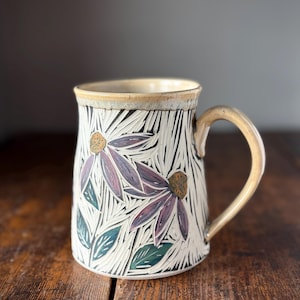 May include: A ceramic mug with a cream-coloured base featuring carved, textured floral patterns. The flowers have purple petals and yellow centres, accented by green leaves. The mug has a smooth, light brown handle and a matching rim.