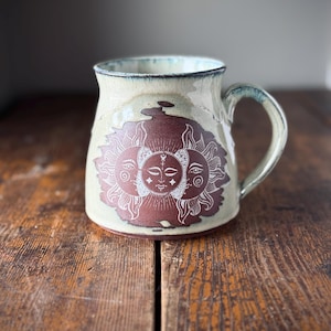Peut inclure: Une tasse en céramique avec une base crème et un motif marron représentant trois visages de soleil. La tasse a un bord et une anse bleus. Le motif est blanc et la tasse est posée sur une surface en bois.