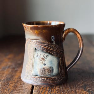 Peut inclure: Une tasse en céramique avec un vernis brun brillant et une anse brun foncé. L'avant présente un cadre en relief texturé entourant une illustration peinte d'un cerf. Le bord et la partie supérieure ont une finition brune lisse et réfléchissante.