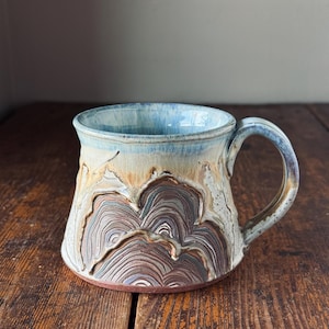 Peut inclure: Une tasse en céramique avec une base large et une anse incurvée. L'extérieur présente un motif en relief sculpté et texturé de formes superposées rappelant des montagnes dans des tons marron et crème, surmonté d'un bord émaillé bleu et blanc.