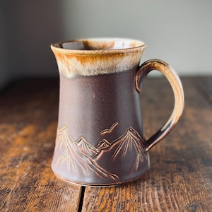 Peut inclure: Une tasse en céramique avec une finition mate brun foncé présentant un motif de chaîne de montagnes en relief et un oiseau en vol. Le bord et l'anse sont recouverts d'un vernis contrastant beige clair et crème, créant un aspect texturé.