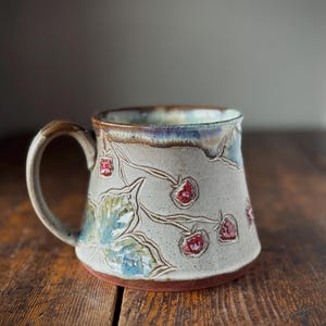 Peut inclure: Une tasse en céramique avec un émail gris clair moucheté, ornée de motifs de fraises rouges en relief et de feuilles vertes. Le bord présente un effet d'émail coulant bleu et brun, avec une anse assortie. La base est de couleur brun rougeâtre.