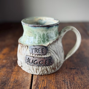 May include: A white ceramic mug with a black handle and the text "But First, Coffee" in a bold, black font. The mug is set against a blurred background, highlighting its clean design.