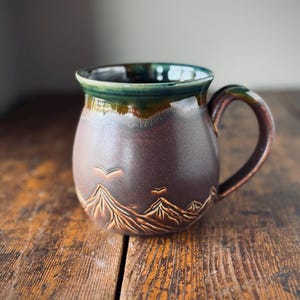 Peut inclure: Une tasse en céramique arrondie avec une finition mate brun foncé présentant des motifs en relief de montagnes et d'oiseaux. Le bord est recouvert d'une glaçure vert profond brillante qui coule sur le corps brun. Elle possède une anse incurvée.